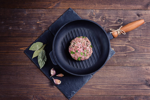 Νωπό βόειο κρέας φέτα με μυρωδικά στο τηγάνι γκριλ σε γκρίζα πέτρα με σκόρδο και φύλλο δάφνης για καφέ φόντο ξύλινη. Ρουστίκ νεκρή. Σκοτεινή, κυκλοθυμική στυλ - Φωτογραφία, εικόνα