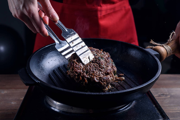 Женщина-повар в красном фартуке переворачивает щипцы хорошо жареного куска мяса на сковородке. темный капризный стиль
 - Фото, изображение