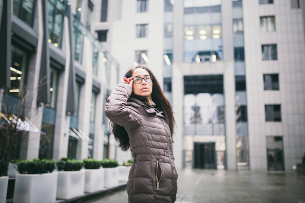 Portrét krásné mladé obchodních žena brunetka v bundu a svetr stojí na pozadí kancelářské budovy, obchodní centrum v brýlích za pohled. Ruka drží na zavěšení sklenic a hledat dál - Fotografie, Obrázek