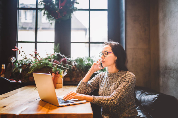 Tema é pequeno negócio. Uma jovem mulher freelance trabalhando atrás de um computador portátil em um café decorado com decoração de Natal e falando ao telefone. Vestido com uma camisola cinza e óculos
 - Foto, Imagem