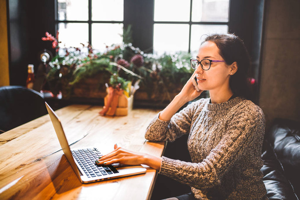 El tema es la pequeña empresa. Una joven independiente trabajando detrás de una computadora portátil en una cafetería decorada con decoración navideña y hablando por teléfono. Vestido con un suéter gris y gafas
 - Foto, imagen