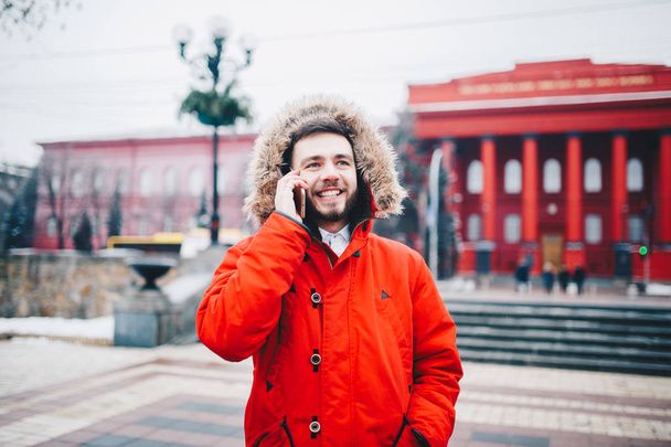 Sakal ve kırmızı ceket başlıklı bir öğrenci genç adam cep telefonu kullanır, üniversite veya kolej bina kırmızı zemin üzerine gülümseme ile telefonda konuşurken yakınındaki baş, elinde tutan - Fotoğraf, Görsel