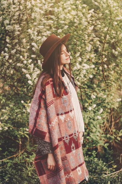 stylish hipster girl in hat and poncho with beautiful hair standing in park in evening sunshine. woman traveler in fashionable outfit relaxing in spring garden. space for text. atmospheric moment - Photo, Image