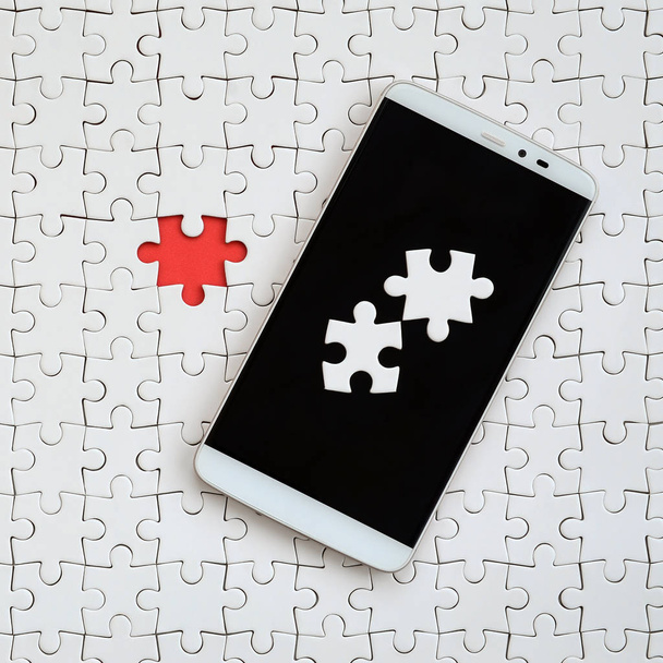 A modern big smartphone with several puzzle elements on the touch screen lies on a white jigsaw puzzle in an assembled state with missing elements - Photo, Image