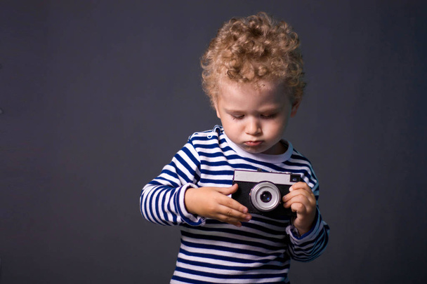 A small curly-haired boy with a camera on a gray background - a studio photo. The kid learns to take pictures.  - Fotografie, Obrázek