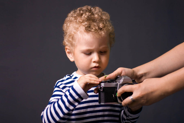 A small curly-haired boy with a camera on a gray background - a studio photo. The kid learns to take pictures.  - 写真・画像