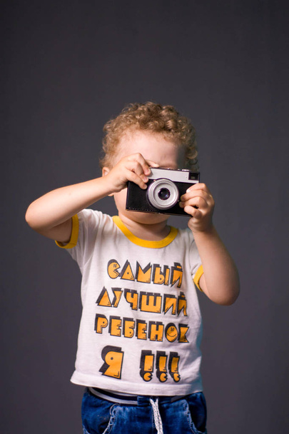 A small curly-haired boy with a camera on a gray background - a studio photo. The kid learns to take pictures.  - Foto, Imagem