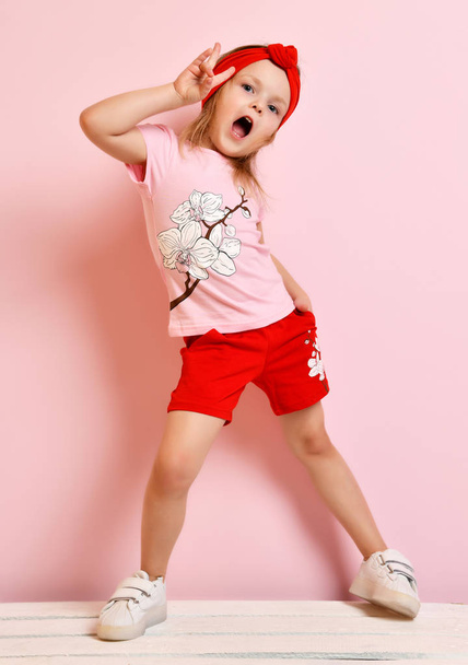 Cute baby girl kid in light pink pajamas and red headband standing posing on pink - 写真・画像