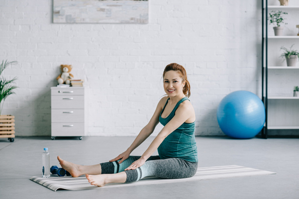 beautiful pregnant woman stretching on yoga mat and looking at camera in living room - Valokuva, kuva