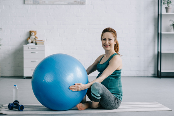 όμορφη έγκυος γυναίκα κάθεται στο χαλάκι γιόγκα με την μπάλα γυμναστικής στο σαλόνι - Φωτογραφία, εικόνα