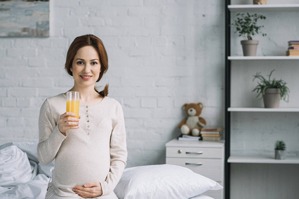 beautiful pregnant woman sitting on bed in bedroom with glass of orange juice - Zdjęcie, obraz