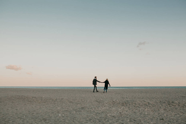 Man and woman walking by the beach sand at sunset, holding hands, with the sea and a blue sky in the background. - Valokuva, kuva
