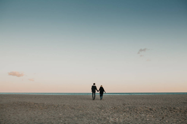 Muž a žena chůze pískem při západu slunce na pláži, držíce se za ruce s mořem a modrá obloha v pozadí. - Fotografie, Obrázek