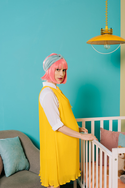 κομψή καρφίτσα έγκυος γυναίκα με ροζ μαλλιά στέκεται κοντά στο παιδικό κρεβατάκι στο δωμάτιο του παιδιού - Φωτογραφία, εικόνα