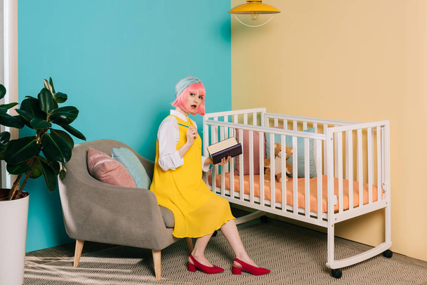 surprised stylish pregnant pin up woman with pink hair holding notebook and looking at camera near baby cot - Photo, Image
