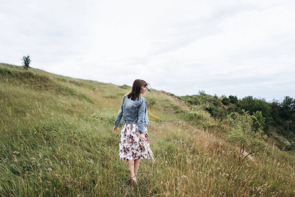 Rückansicht einer Frau in Jeansjacke und Blumenrock, die auf der Wiese spaziert - Foto, Bild