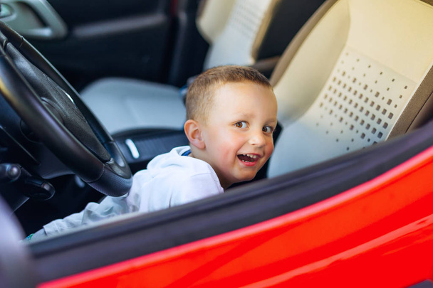 mały chłopiec bawi się na przednim siedzeniu czerwonego samochodu i uśmiecha się szczerze - Zdjęcie, obraz