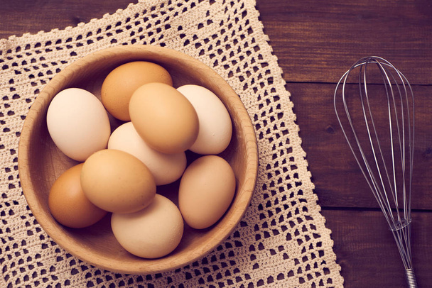 Top view of fresh raw chicken eggs in wooden bowl with napkin and whisk on rustic wooden background. - Fotoğraf, Görsel