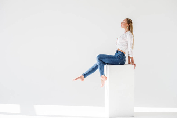 Portrait of sexy blonde girl wearing blue jeans and white shirt sitting on white studio background - Photo, image