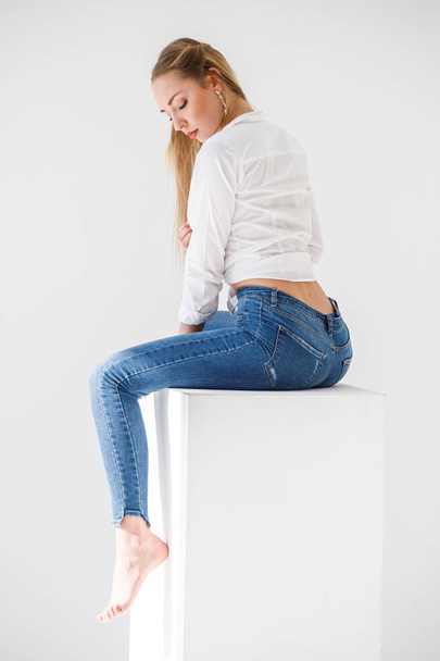 Portrait of sexy blonde girl wearing blue jeans and white shirt sitting on white studio background - 写真・画像
