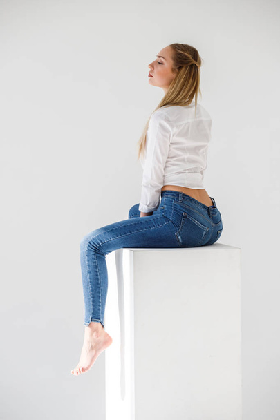 Portrait of sexy blonde girl wearing blue jeans and white shirt sitting on white studio background - Foto, Imagen