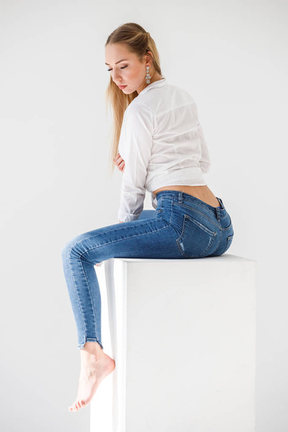 Portrait of sexy blonde girl wearing blue jeans and white shirt sitting on white studio background - Photo, image