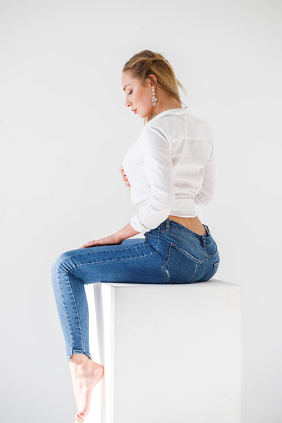 Portrait of sexy blonde girl wearing blue jeans and white shirt sitting on white studio background - Foto, Imagem
