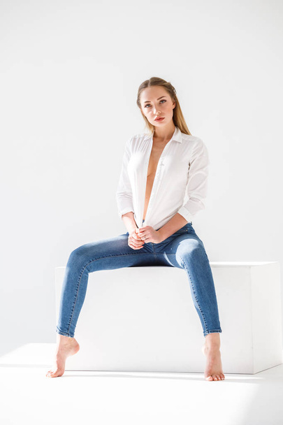 Portrait of sexy blonde girl wearing blue jeans and white shirt sitting on white studio background - Photo, Image