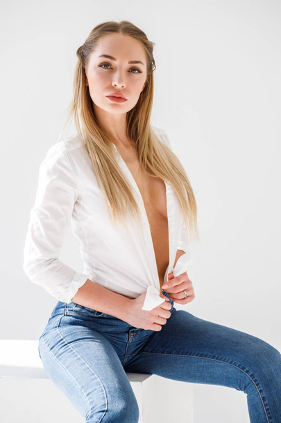 Portrait of sexy blonde girl wearing blue jeans and white shirt sitting on white studio background - Fotografie, Obrázek