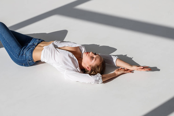 Portrait of sexy blonde girl wearing blue jeans and white shirt lying on white studio background - Fotografie, Obrázek