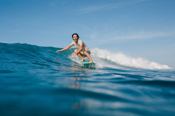 glücklicher junger Mann im nassen T-Shirt, der an sonnigen Tagen Wellen auf dem Surfbrett reitet - Foto, Bild
