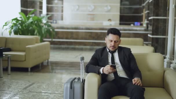 Pan shot of concentrated businessman using smartphone sitting on armchair inside luxury hotel with luggage near him - Filmati, video