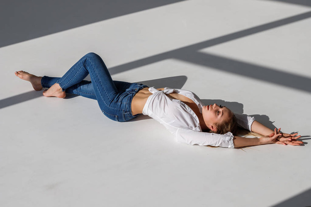 Portrait of sexy blonde girl wearing blue jeans and white shirt lying on white studio background - Фото, изображение