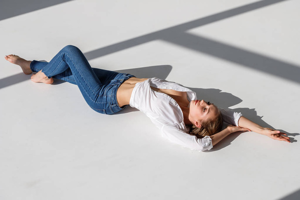 Portrait of sexy blonde girl wearing blue jeans and white shirt lying on white studio background - Фото, зображення