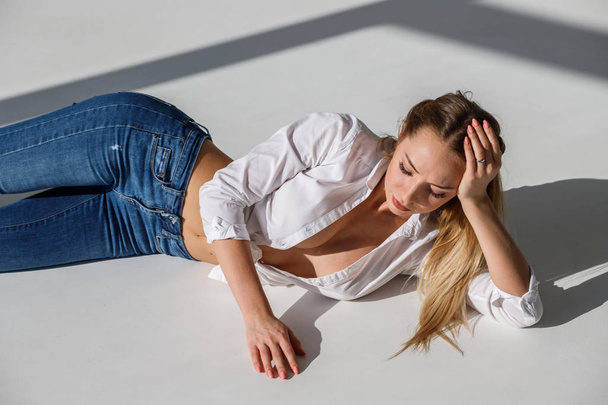 Portrait of sexy blonde girl wearing blue jeans and white shirt lying on white studio background - Fotografie, Obrázek