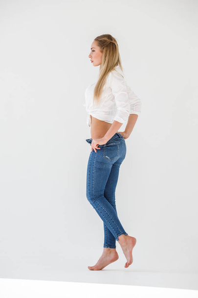 Portrait of sexy blonde girl wearing blue jeans and white shirt standing on white studio background - Фото, изображение