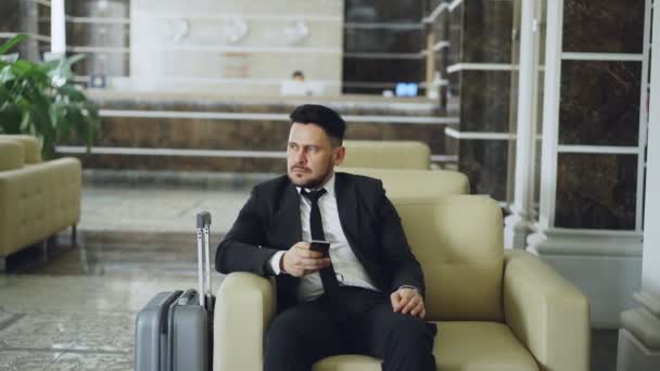 Pan shot of thoughtful businessman looking aside and using smartphone sitting on armchair in luxury hotel with luggage near him. Travel, business and people concept - Кадры, видео