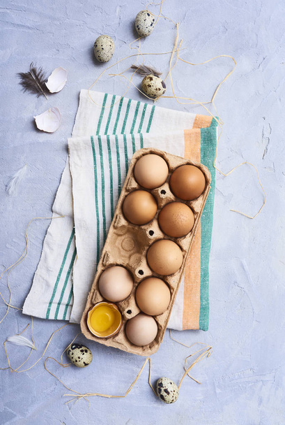 Свіжі сирі курячі яйця в картонній коробці на кухонному рушнику та перепелині яйця з пір'ям на синьому бетонному фоні
. - Фото, зображення