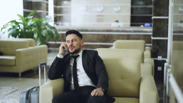 Pan shot of confident businessman talking mobile phone while sits on armchair in luxury hotel with luggage near him. Travel, business and people concept - Filmati, video