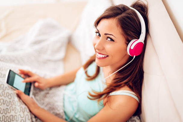 smiling woman relaxing in bed and listening to music from smartphone on headphones - Foto, imagen