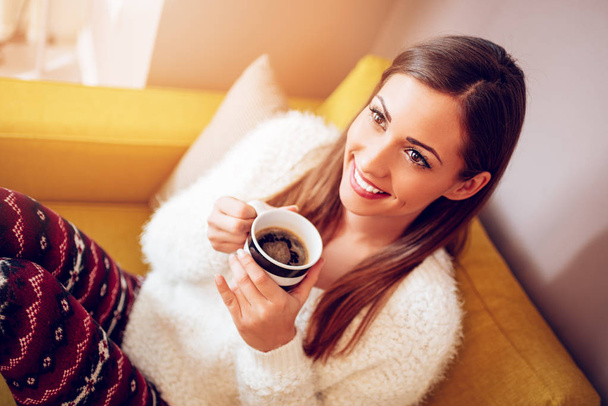 молода брюнетка сидить на дивані у вітальні і п'є каву
 - Фото, зображення