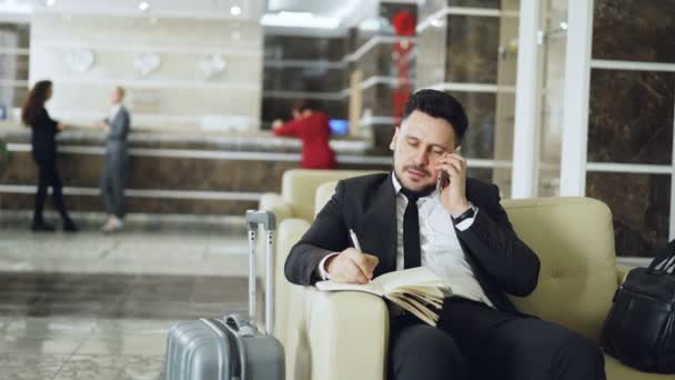 Pan shot of confident businessman talking mobile phone and writing notes in notepad while sits on armchair in luxury hotel with luggage near him. Travel, business and people concept - Footage, Video