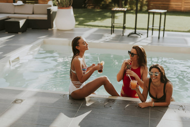 Молоді жінки п'ють коктейлі і розважаються біля басейну в спекотний літній день
 - Фото, зображення