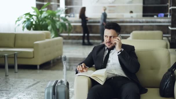 Pan shot of cheerful confident businessman talking mobile phone smiling and writing notes in notepad while sits on armchair in hotel reception. Travel, business and people concept - Záběry, video