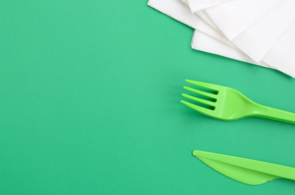Disposable plastic cutlery green. Plastic fork and knife lie on a green background surface next to napkins - Photo, Image