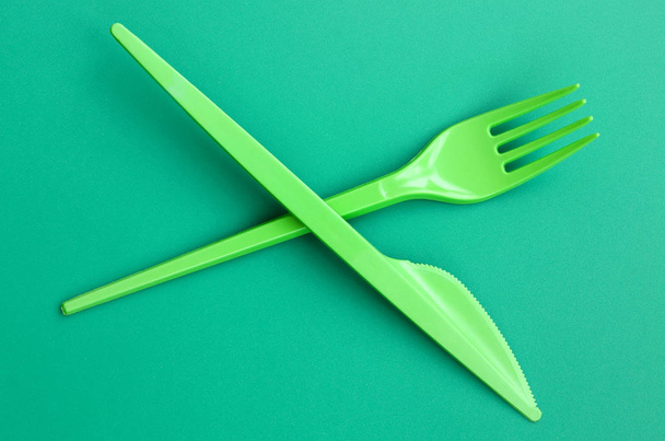 使い捨てのプラスチック製食器グリーン。緑の背景の表面に横にプラスチックのフォークとナイフ - 写真・画像
