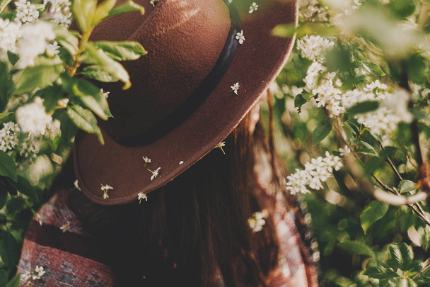 beautiful white flowers on hat of hipster girl in green leaves with bloom in evening sunshine. stylish woman traveler calm relax in spring garden - Fotoğraf, Görsel