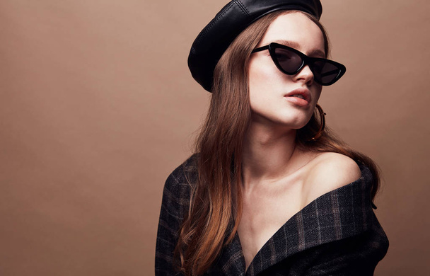 Fashion portrait of beautiful young woman in black leather beret cap, plaid jacket and cat eye retro sunglasses with massive golden earrings - Foto, Imagem