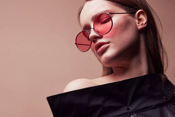 Warm colored fashion portrait of beautiful young woman in round red sunglasses and grey latex jacket posing in studio - Valokuva, kuva