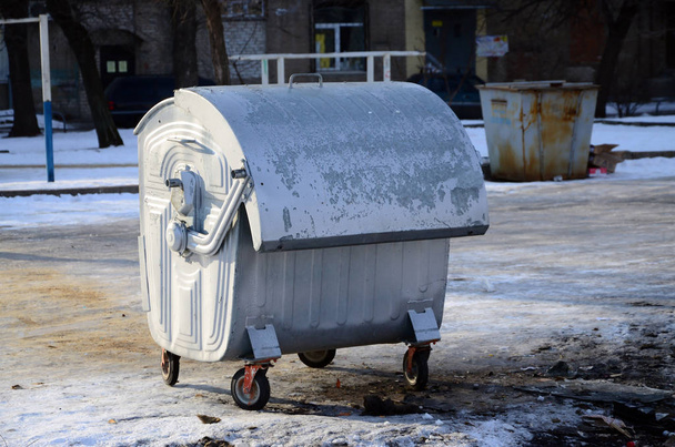 Um recipiente de lixo prateado fica perto de edifícios residenciais no inverno
 - Foto, Imagem